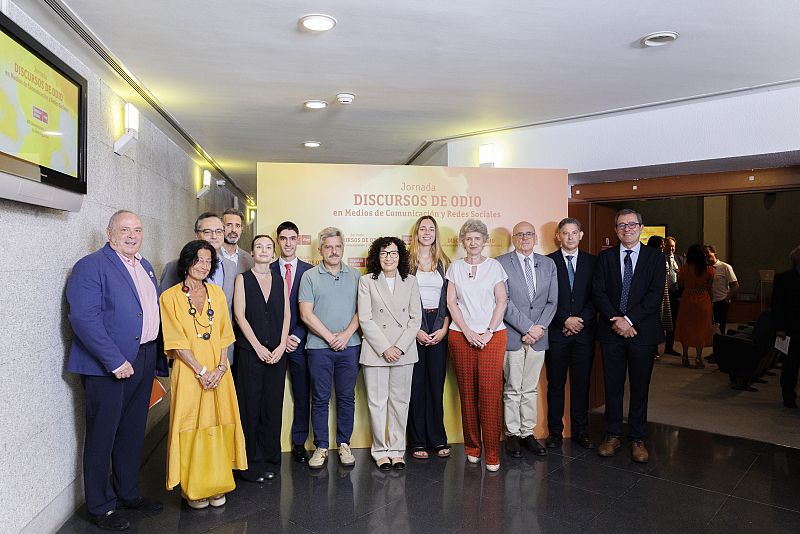 Grupo de personas en evento sobre discursos de odio en medios y redes sociales. Cartel y pantalla muestran el título del evento.