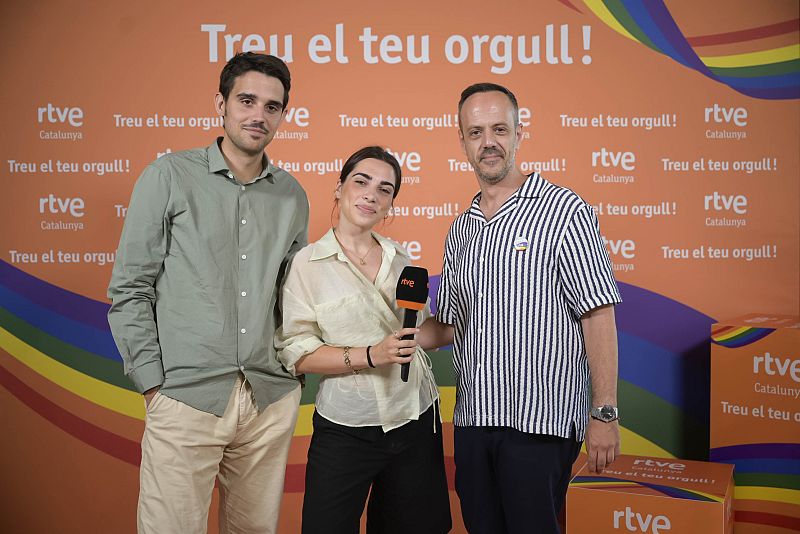 Foto promocional con tres personas (dos hombres y una mujer) frente a un fondo naranja con arcoíris y el lema "Treu el teu orgull!". La mujer porta un micrófono con logo RTVE; cajas con el mismo logo aparecen en la esquina inferior derecha.