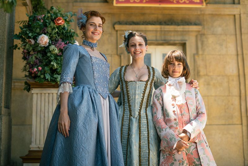 Tres personas en atuendos del siglo XVIII posan para una foto frente a un edificio clásico.  Una mujer sonríe, otra mira a la cámara y un joven también posa.  Un arreglo floral está presente.