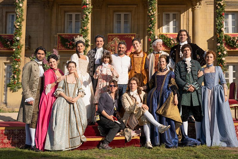 Actores en trajes de época del siglo XVIII posan en una tarima con alfombra roja frente a un edificio. Una persona con ropa moderna está presente, posiblemente del equipo de producción.
