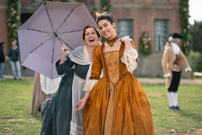 Rodaje de serie: dos mujeres en trajes de época, una con vestido gris y delantal, la otra con vestido mostaza, ríen animadamente en un exterior soleado con un edificio decorado con motivos navideños al fondo.