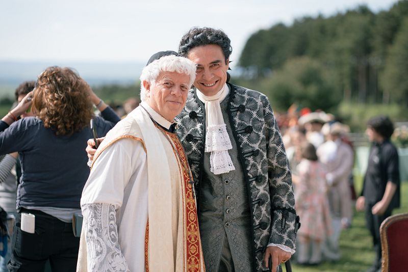 Actores en rodaje de época: sacerdote y hombre elegante sonríen a cámara. Fondo con equipo y extras, espacio exterior arbolado.