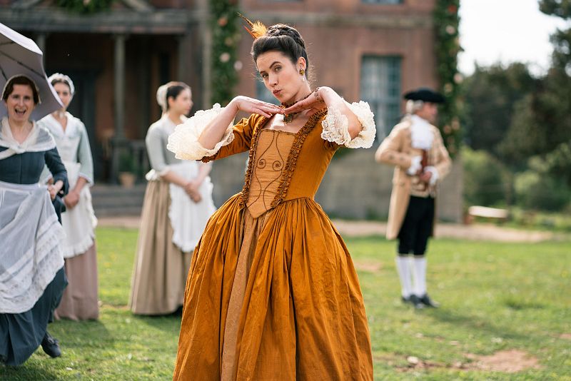 Mujer con vestido ocre y tocado de plumas posa en jardín con otras mujeres y un hombre con peluca, en una escena que sugiere una serie de época.