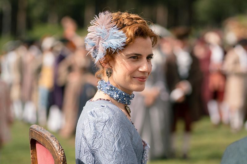 Mujer con peinado trenzado y tocado de plumas, traje azul de época, sonríe ligeramente. Fondo con figuras vestidas de época.