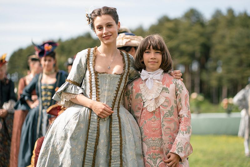 Mujer y niño/a con atuendos de época (siglo XVIII) en un jardín.  Imagen de una producción, posiblemente el making-of de una serie.