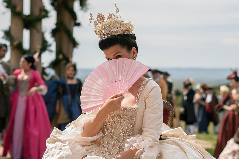 Mujer con vestido crema y corona, sentada con un abanico, rodeada de otras personas en atuendos de época.  Ambiente de fiesta aristocrática.