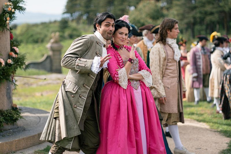 Pareja en trajes de época (siglo XVIII) posando alegremente para la cámara durante un rodaje al aire libre. Fondo con arquitectura de piedra y rejas.