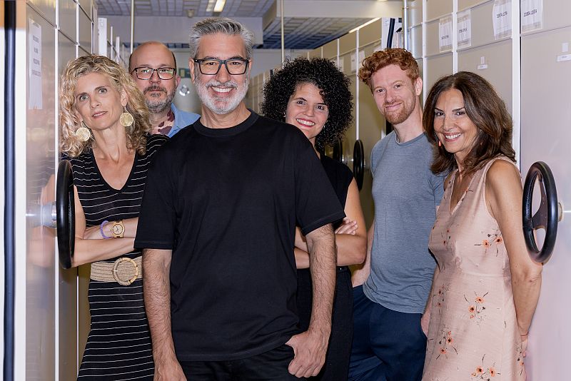 Grupo de seis personas (tres hombres y tres mujeres) sonrientes, posando en un archivo con estanterías metálicas.  Vestimenta variada.
