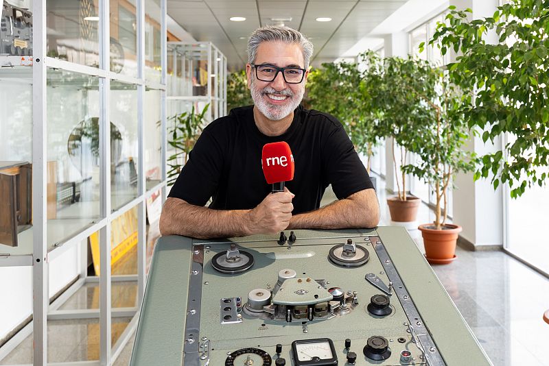 Carles Mesa, con camiseta negra y gafas, sonríe sujetando un micrófono de RNE en un estudio de radio con aparatos antiguos y plantas.