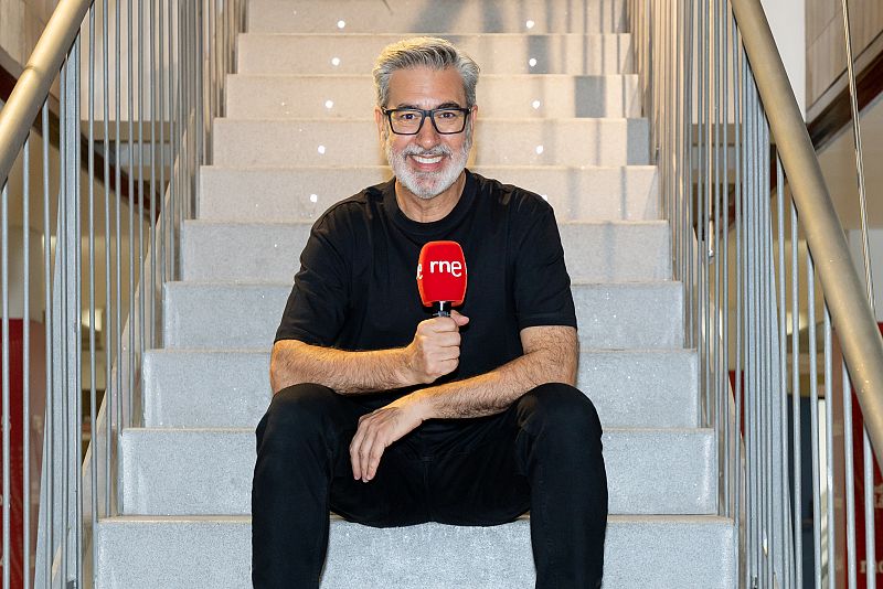 Presentador de radio con micrófono RNE, sentado en escaleras de cemento, viste camiseta y pantalones negros, sonríe a cámara.