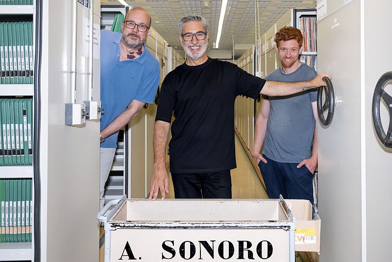 Tres hombres trabajan en un archivo con estanterías y un carrito marcado 'A. SONORO'. Sonríen a la cámara, mostrando una escena de trabajo en un almacén de archivos.