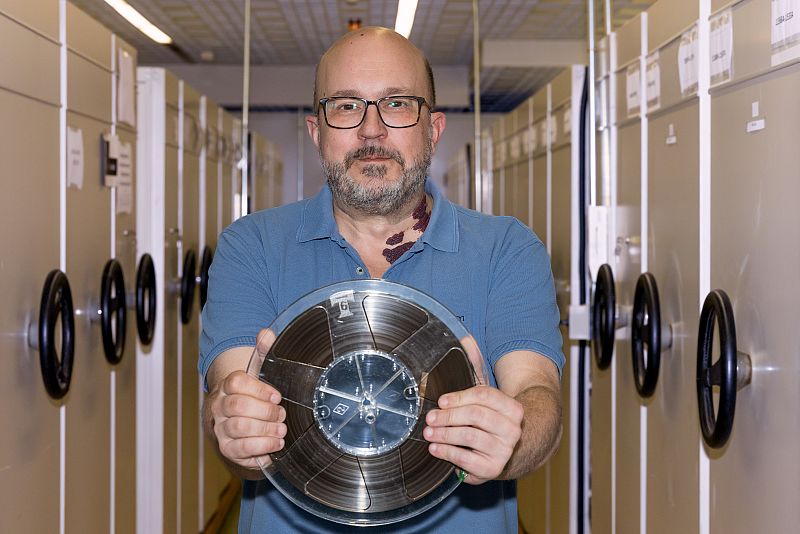 Hombre con barba y gafas sostiene una cinta magnética en un archivo con estanterías metálicas.  Posible trabajo de archivo y preservación de grabaciones.