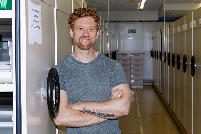 Hombre pelirrojo con barba y tatuaje en el antebrazo, apoyado en estantería beige con cartel "ORIGINALISTAS. RECOGIDA. PROTECCIÓN. FONDO SONORO".