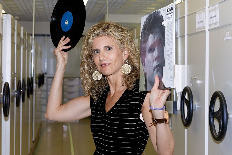 Mujer con blusa a rayas sostiene un vinilo y portada con el nombre "JOSE MANUEL". Sonríe a cámara, en pasillo con estanterías metálicas. Lleva pendientes dorados y pulsera morada.