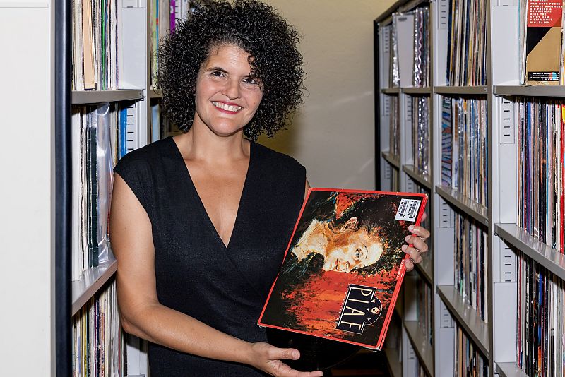 Mujer sonriente sosteniendo un disco de vinilo con portada roja y naranja;  el nombre "PIAF" es visible.  Entorno con estanterías llenas de objetos similares.