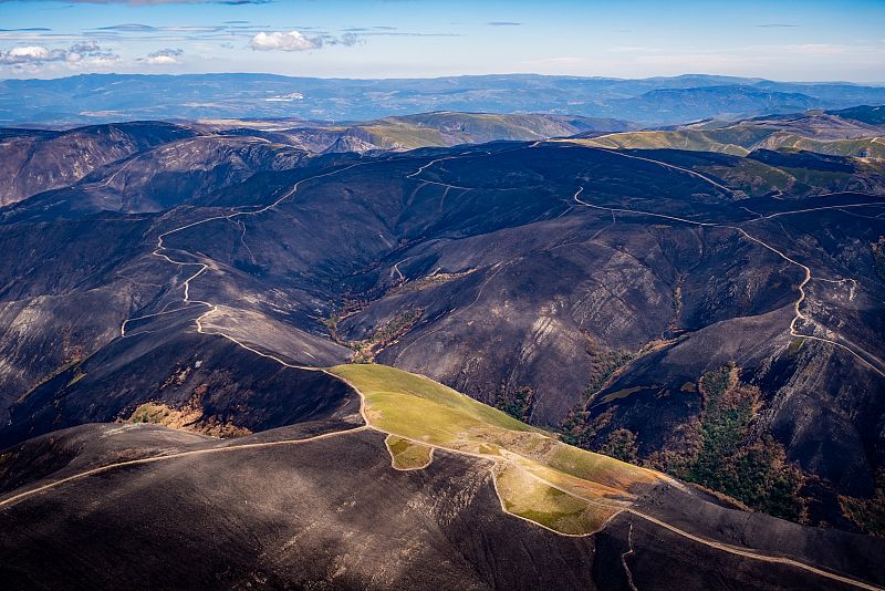 Vista aérea de un incendio en zona montañosa, mostrando un paisaje parcialmente quemado con carreteras que atraviesan el terreno.