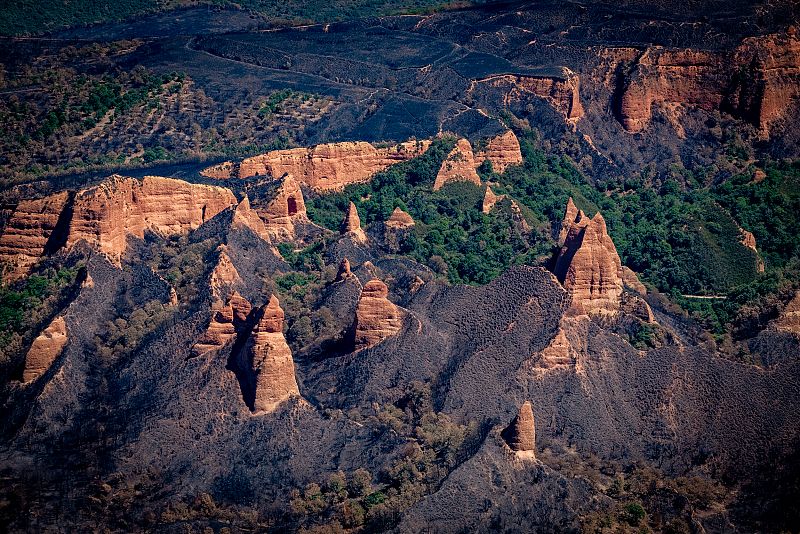 Vista aérea de Las Médulas mostrando la devastación de un incendio.  Tierra quemada de color oscuro contrasta con las formaciones rocosas rojizas y zonas verdes no afectadas.