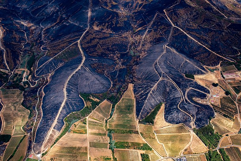 Secuelas de un incendio forestal en zona montañosa: extensas áreas quemadas, contrastando con vegetación y cultivos supervivientes.  Se aprecian cortafuegos y pequeñas edificaciones.