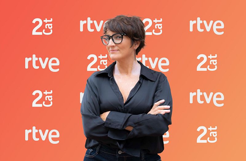 Silvia Abril, con gafas y pendientes, posa con una camisa negra y vaqueros frente a un fondo naranja con logotipos repetidos.