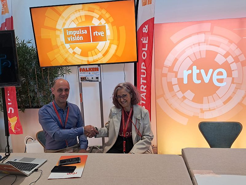 Dos personas, un hombre y una mujer, se dan la mano sobre una mesa. El hombre lleva una camisa azul y la mujer gafas, camiseta negra y chaqueta crema. Se observa una pantalla con logos y un banner publicitario.
