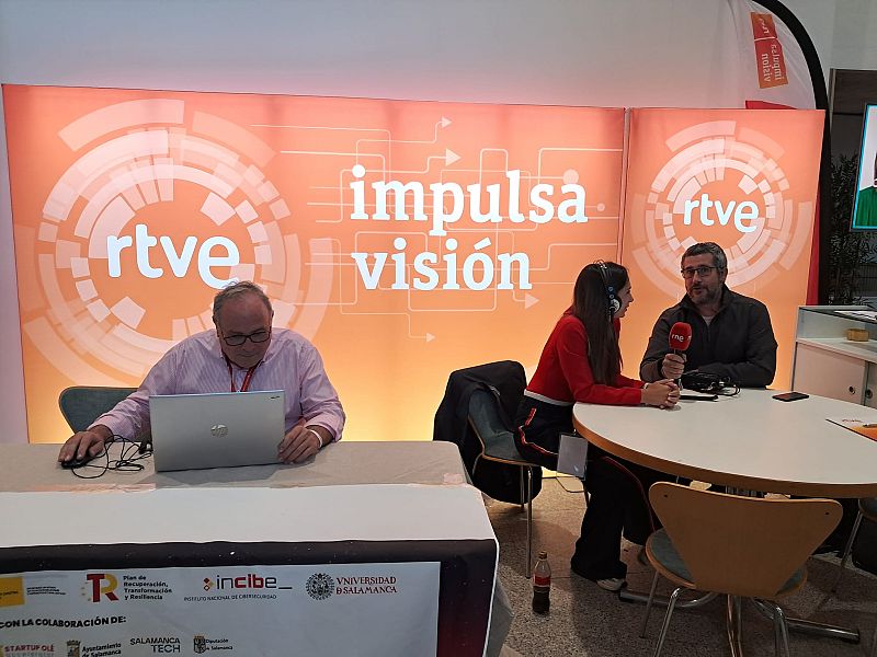 En un stand, se observa a dos personas, un hombre y una mujer, interactuando. La mujer, con auriculares, entrevista a un hombre con barba. Un ordenador portátil y un micrófono son visibles, junto a logotipos de varias organizaciones.