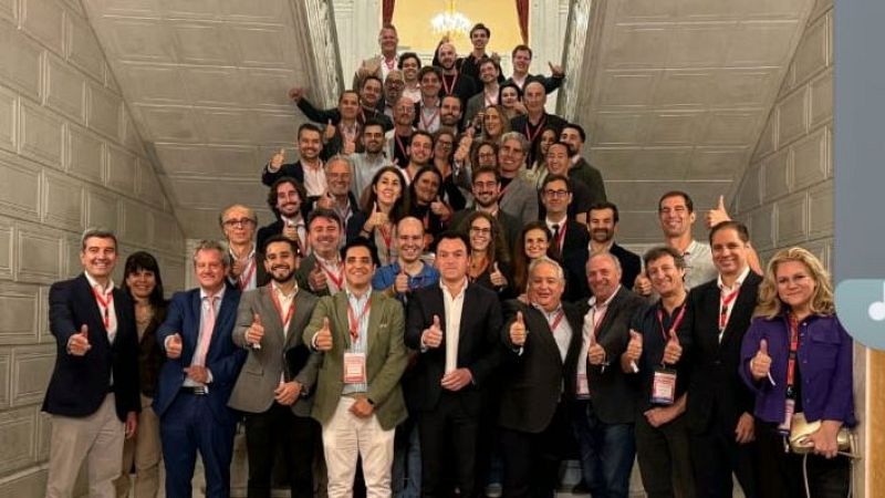 En la escalinata de un edificio, un grupo numeroso de personas sonríe y levanta el pulgar, mostrando acreditaciones colgadas al cuello.