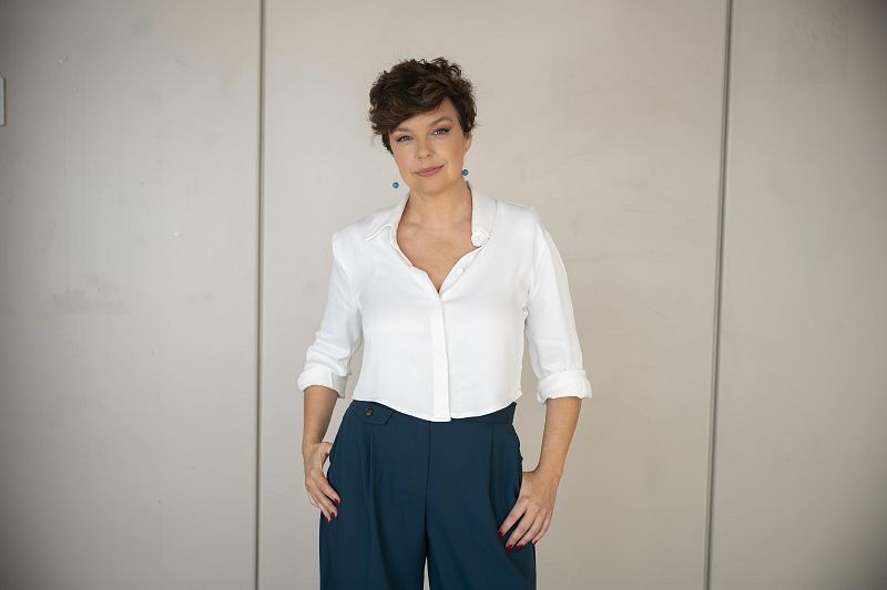 Cristina Villanueva, con el pelo castaño y peinado moderno, posa frente a una pared gris. Viste una camisa blanca y pantalones oscuros, con pendientes turquesa y las manos en los bolsillos.