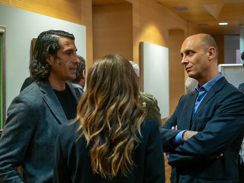 En un espacio interior, tres personas interactúan: un hombre con chaqueta gris conversa con una mujer de espaldas, mientras que un hombre con traje azul observa. Se aprecia la presencia de una persona con uniforme militar.