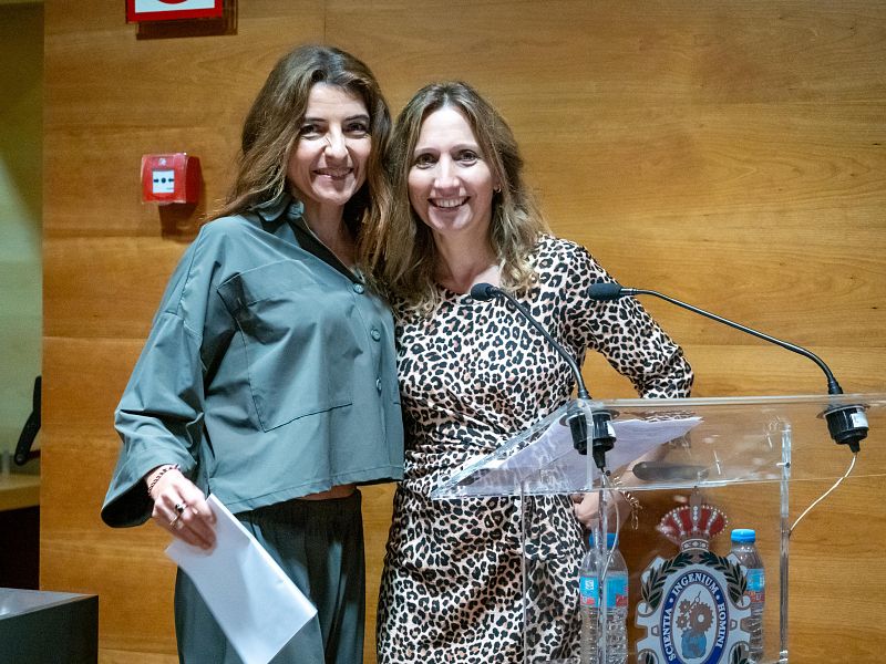 En un evento, dos mujeres posan sonrientes. Una de ellas, con vestimenta oscura, sostiene un papel, mientras que la otra, con un vestido estampado, se encuentra detrás de un atril con micrófonos y el logo de una empresa.