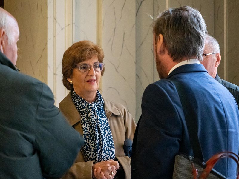 En un interior con diseño de mármol, una mujer con gafas y chaqueta beige conversa con otros individuos, algunos de espaldas, que visten trajes oscuros. La mujer lleva una bufanda estampada y su expresión es amigable.
