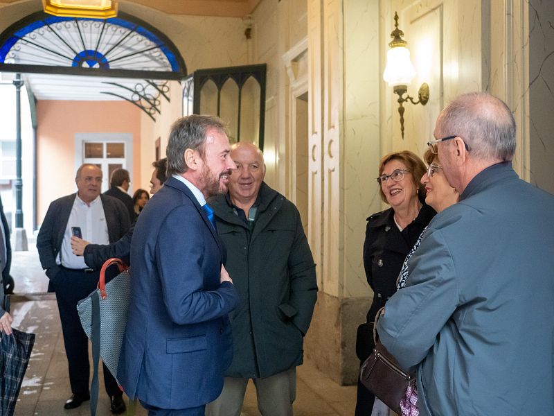 En un espacio interior con elementos arquitectónicos clásicos, un grupo de personas conversan. Se distinguen hombres con trajes y chaquetas, y una mujer con gafas y abrigo negro. El evento parece estar relacionado con "Impulsa Visión".