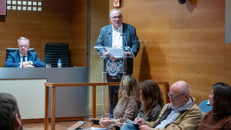 En una sala de conferencias, un hombre con gafas y pelo canoso, vestido con camisa blanca y chaqueta gris, habla desde un atril transparente. Detrás, un hombre en traje azul escucha, y la audiencia presta atención a la presentación.