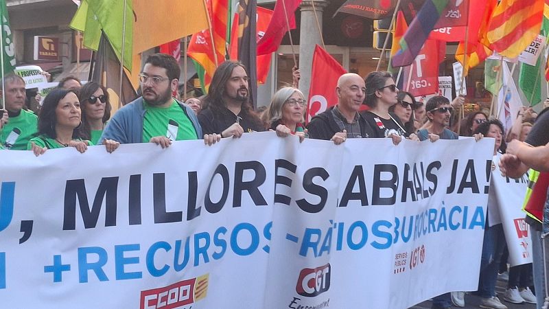 Una multitud de persones, possiblement professors, es manifesta a Barcelona amb una pancarta que demana millores i més recursos. Es distingeixen banderes de diversos colors i logotips sindicals.