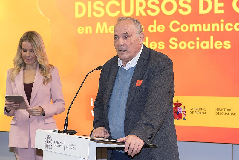 En un evento, Esteban Mayoral, con camisa azul y jersey gris, habla desde un atril con logotipos gubernamentales. Una mujer rubia con chaqueta rosa, a su lado, sostiene una tablet.