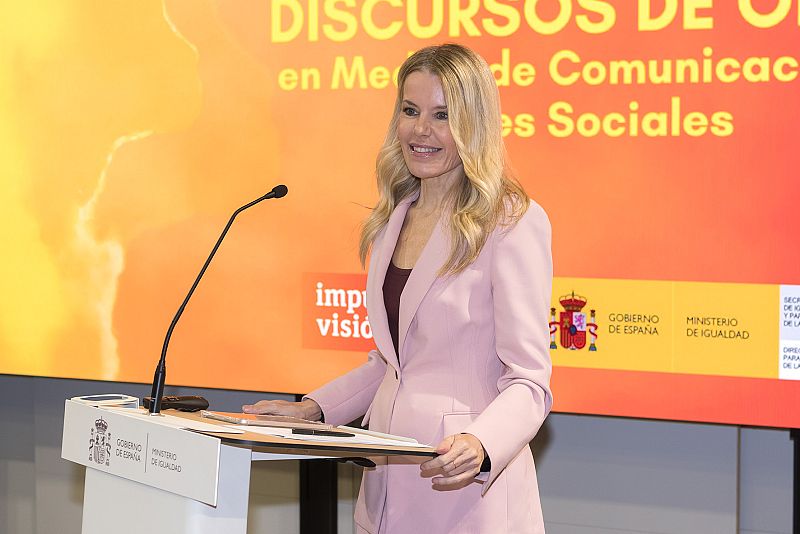 Una mujer rubia, vestida con un traje rosa claro, se encuentra frente a un atril blanco con el escudo de España y el logo de un ministerio, sonriendo mientras un micrófono está colocado frente a ella.