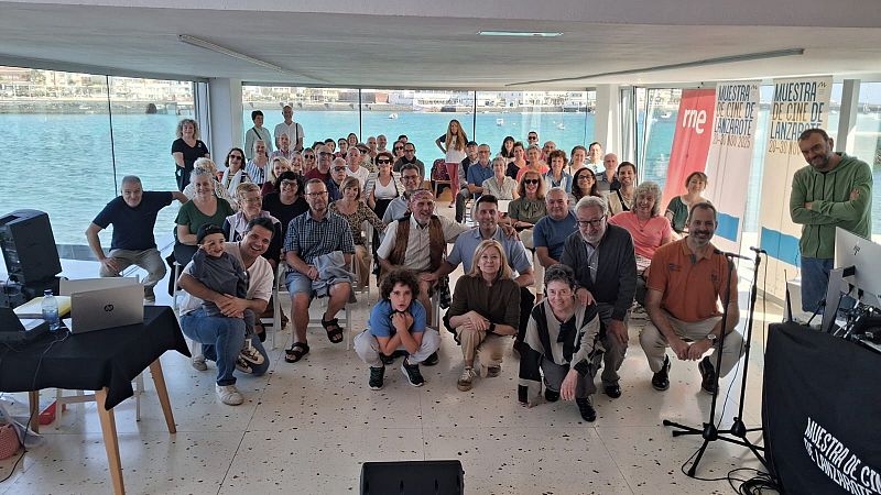 Un grupo de personas de diferentes edades se reúne en un espacio interior con vistas al mar, algunas sentadas y otras de pie. Hay dos mesas con equipos de sonido y un portátil, y al fondo se ven carteles promocionando un evento con fechas específicas.