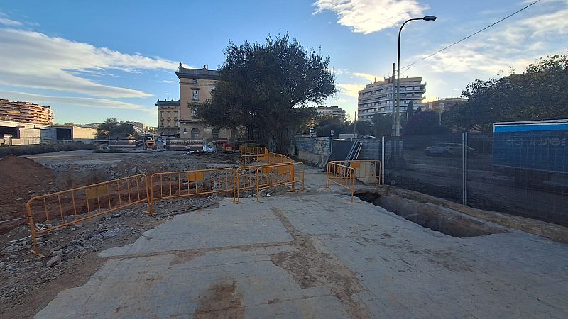 Una zona en construcción con una excavación y barreras de seguridad naranjas se encuentra frente a un edificio de varios pisos, posiblemente una estación, con un árbol cercano y un cielo nublado.