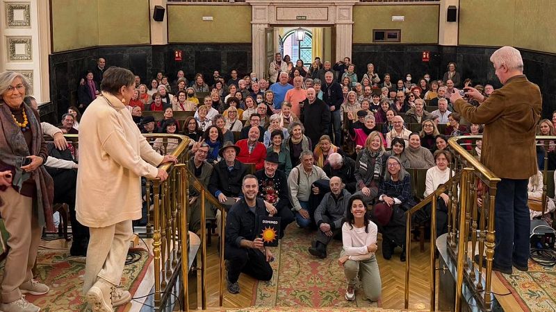 Un numeroso grupo de personas se reúne en un salón de actos, posiblemente en Madrid, el 14 de diciembre. Se observa a una mujer de pie y a un hombre tomando fotos, mientras otros están sentados o arrodillados en las escaleras. Un cartel con una palabra y un sol es visible.