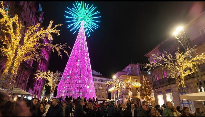 En la Porta do Sol, un gran árbol de Navidad de luces rosas y una estrella en la cima atrae a una multitud. El ambiente festivo se completa con la iluminación de los edificios y árboles más pequeños, creando una escena navideña.