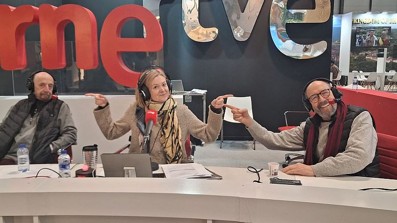 Tres personas, dos hombres y una mujer, sonríen y señalan algo mientras están sentadas en una mesa con micrófonos y ordenadores, en un stand con los logos de dos cadenas de televisión y radio.