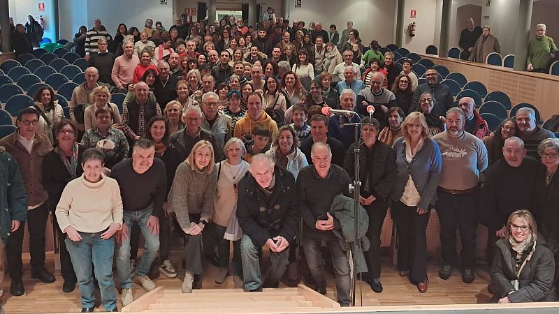 En un auditorio, un grupo diverso de personas posa para una foto. Se observan butacas azules y una escalera, con un fondo de paredes claras.