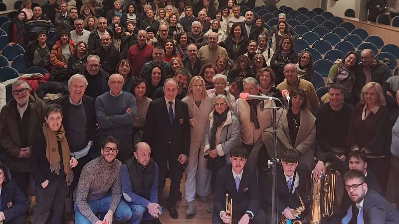 Un evento en un auditorio con un grupo de músicos jóvenes tocando instrumentos musicales. Se distinguen personas vestidas formalmente, incluyendo un hombre con una insignia, y una audiencia numerosa en las butacas.