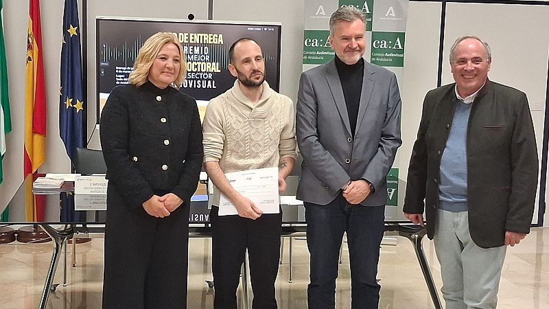 En una ceremonia, un hombre joven sostiene un certificado, flanqueado por otras tres personas. Detrás de ellos, una pantalla anuncia el premio y los logos de las instituciones patrocinadoras, con banderas de España y la Unión Europea a un lado.
