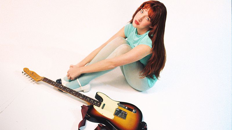 Una joven pelirroja, con una camiseta aguamarina y zapatos blancos, está sentada en el suelo con las piernas cruzadas, mirando hacia arriba. A su lado, se encuentra una guitarra eléctrica.