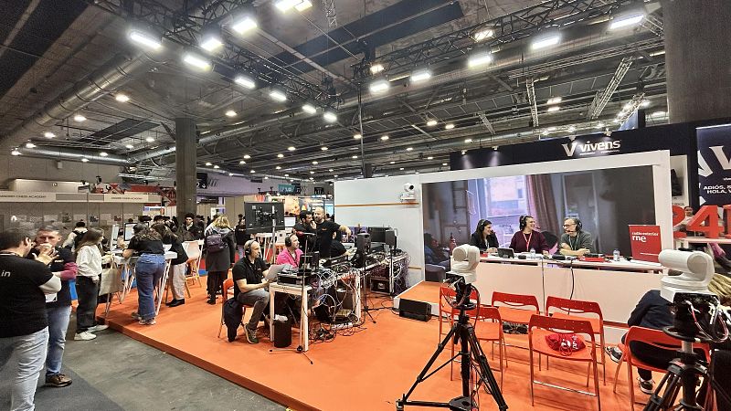 En un recinto ferial, un set de grabación con cámaras y micrófonos es el centro de atención. Tres personas están sentadas a una mesa con el logo "radio marca me", posiblemente grabando un programa. Otros stands y personas interactuando completan la escena.