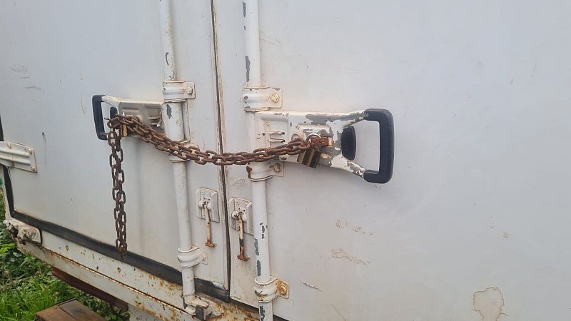 Una puerta metálica blanca, con señales de óxido, está cerrada con una cadena y un candado oxidados, que aseguran los tiradores.