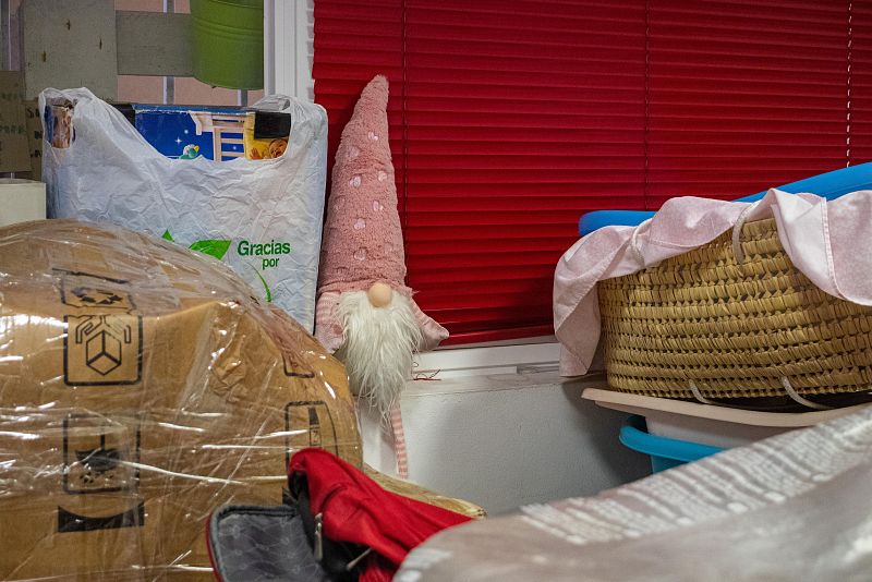 Una caja de cartón envuelta en plástico, una bolsa con texto, un gnomo de peluche con gorro rosa y barba blanca, y una cesta de mimbre con tela blanca, componen la imagen, junto a recipientes de plástico y una ventana con persiana roja.