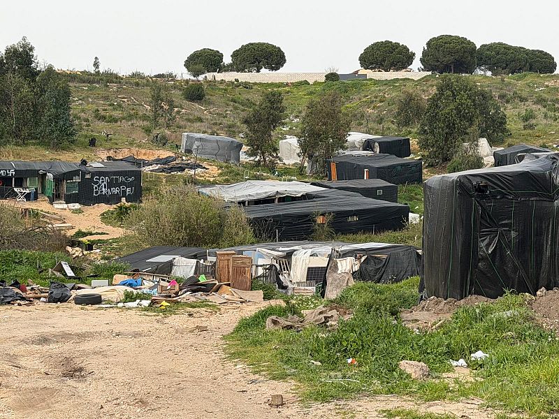 Un asentamiento chabolista con construcciones precarias y cubiertas oscuras se extiende sobre un terreno irregular. Se observa la inscripción "SANTIAGO BERNABEU" en una de las estructuras, en un entorno descuidado y con cielo nublado.