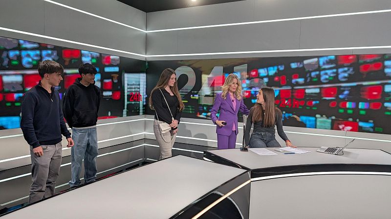 En un estudio televisivo, una mujer rubia vestida de lila interactúa con una joven sentada frente a un ordenador portátil. Dos jóvenes están de pie a los lados y otra junto a la joven sentada. El fondo muestra pantallas con contenido diverso.