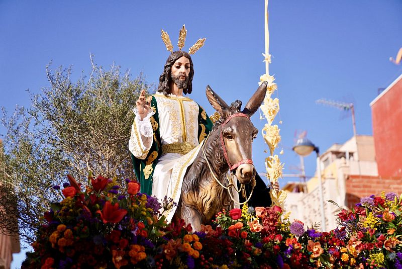 Una estatua de Jesús, con una túnica blanca y detalles dorados, montado sobre un burro marrón con un arnés rojo, rodeado de flores coloridas y con un cielo azul de fondo.